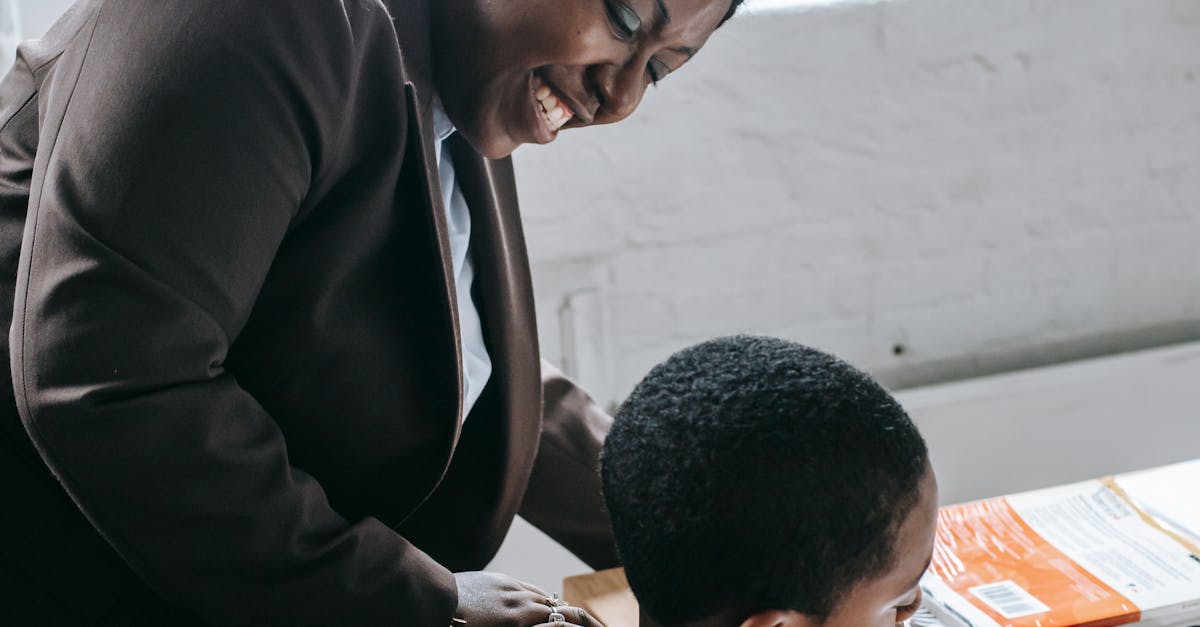 High angle of smiling adult ethnic female tutor teaching attentive little boy solving task during lesson in classroom
