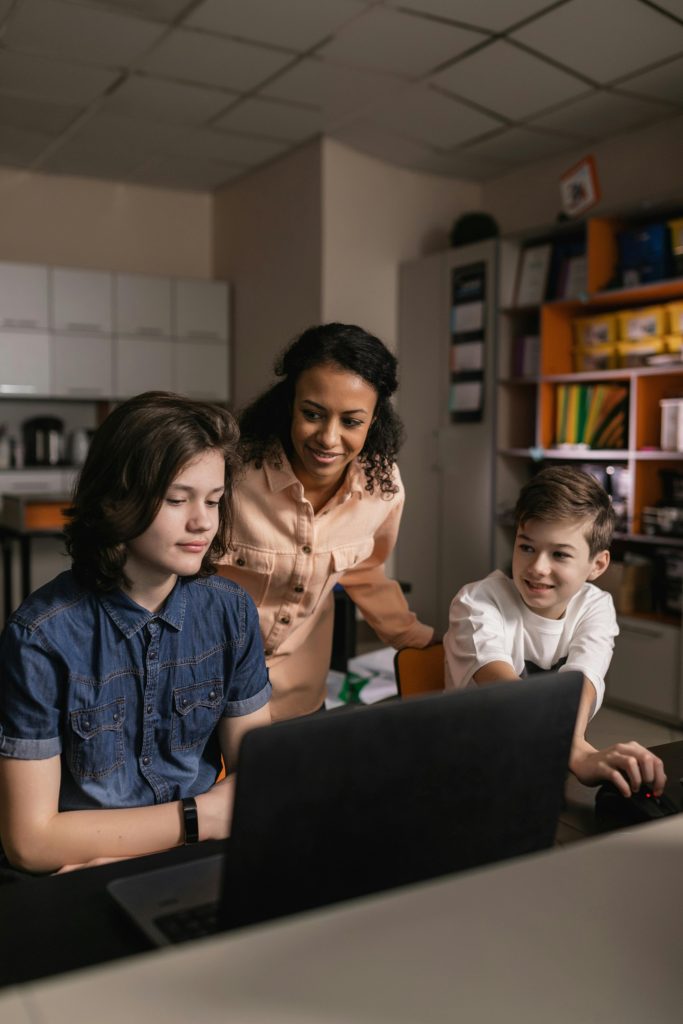 Teacher assisting diverse students in a classroom with a laptop, promoting teamwork and education.
